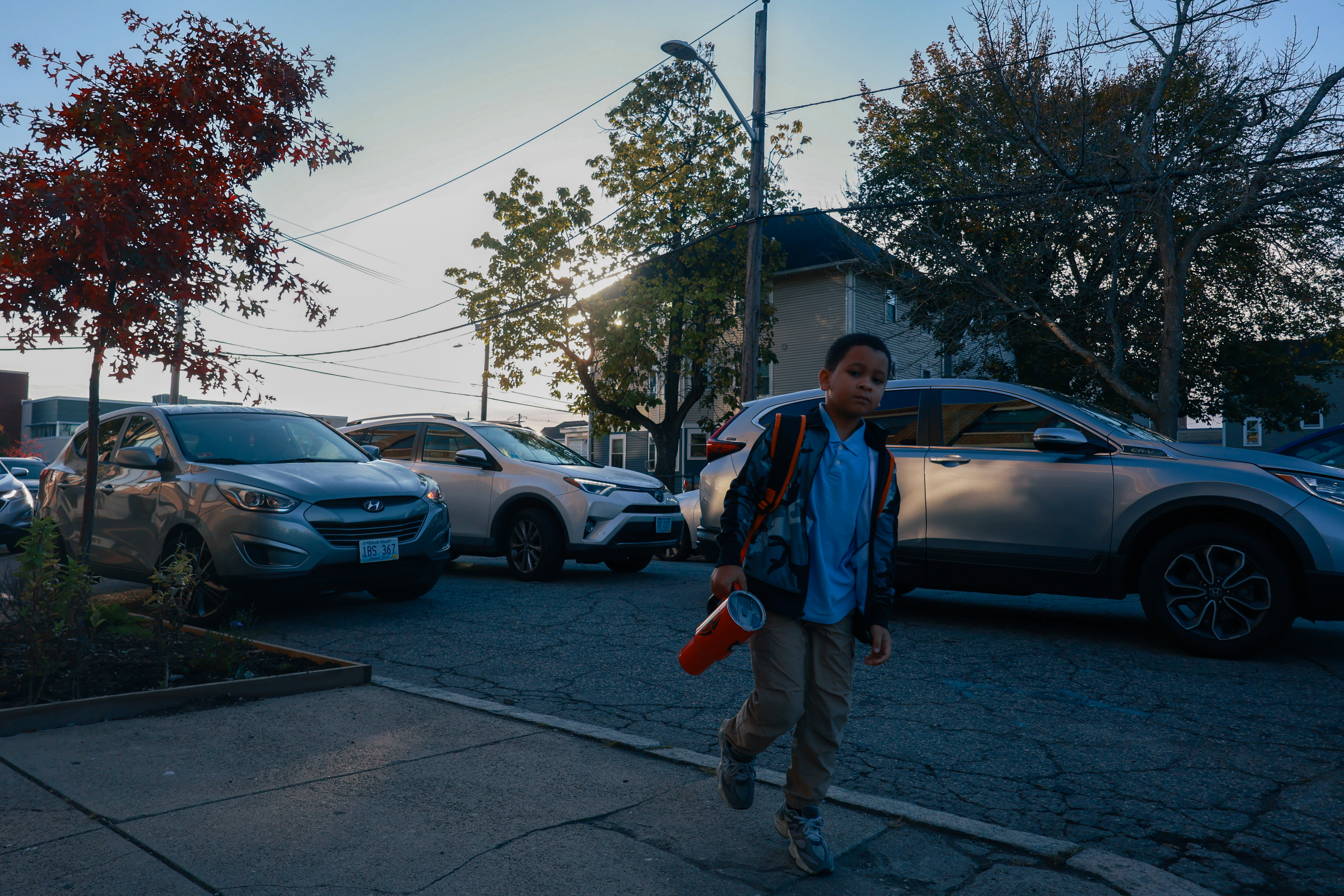 Photo of a young student walking towards the viewer holding a large red thermos and wearing a backpack, with cars lit by the sunrise on the street behind him.
